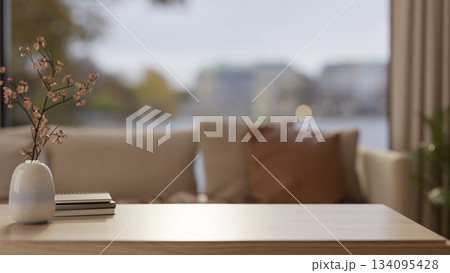 Flowering branch in ceramic vase and books on wooden table across sofa and glass wall of living room 134095428
