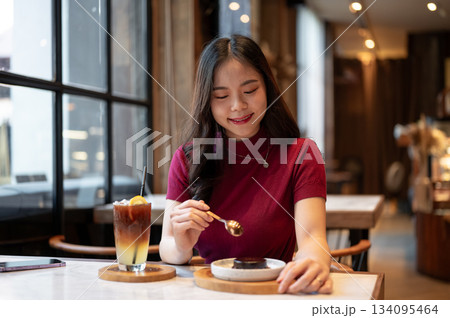 Young asian woman holding spoon eating chocolate cake aside orange coffee cup at table in the cafe. 134095464