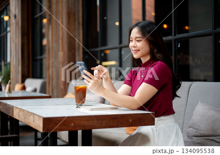 Smiling asian woman looking and pointing at phone aside coffee while sitting on sofa at cafe's table 134095538