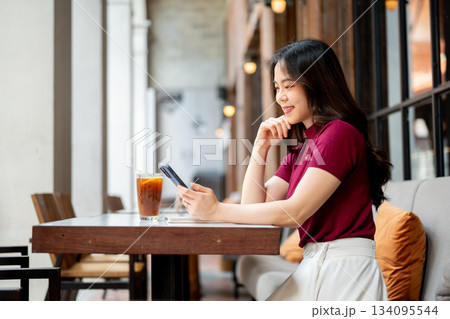 Asian woman holding and looking at phone aside coffee on table while sitting on sofa in the cafe. 134095544
