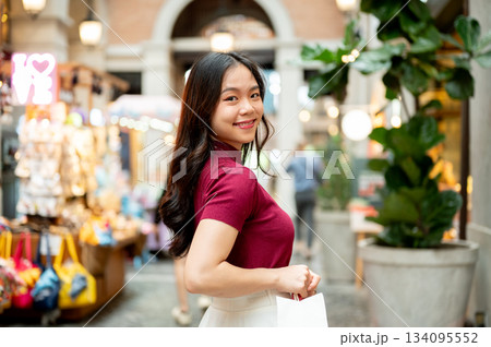 Smiling asian woman holding a paper shopping bag while standing sideway in an indoor market or mall. Smiling asian woman holding a paper shopping bag while standing sideway in an indoor market or mall. 134095552