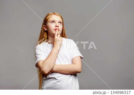 Thoughtful teenage girl with long golden blond hair looking up in studio, isolated on gray background, young female model in white clothing posing Thoughtful teenage girl with long golden blond hair looking up in studio, isolated on gray background, young female model in white clothing posing 134096317