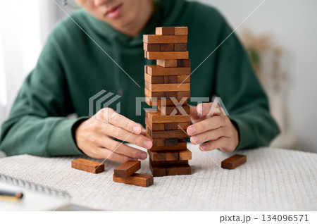 A man looking and hand taking out a piece of wooden block in tower building game while playing alone 134096571