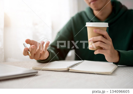 Close up of a man holding coffee cup and pen over notebook aside laptop sitting at table in a cafe. Close up of a man holding coffee cup and pen over notebook aside laptop sitting at table in a cafe. 134096605