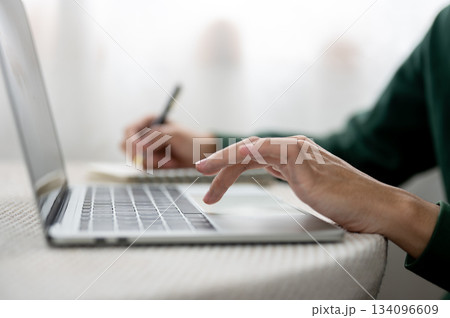 Close up of hands holding pen writing in notebook and typing on laptop as sitting at table in a cafe Close up of hands holding pen writing in notebook and typing on laptop as sitting at table in a cafe 134096609
