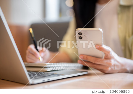 Close up woman student holding pen writing in notebook looking at phone aside laptop on table in cafe 134096896