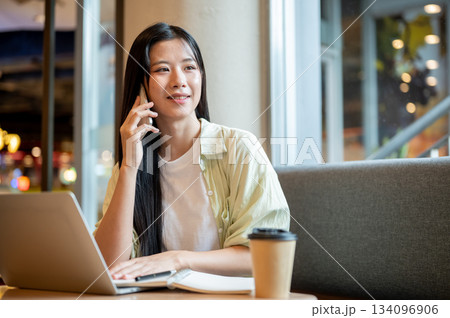 Asian woman student holding and talking on phone over notebook and laptop on wooden table in a cafe. Asian woman student holding and talking on phone over notebook and laptop on wooden table in a cafe. 134096906