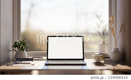 Blank screen laptop with calculator and coffee cup on wooden table under window in warm sunlight. 134096916