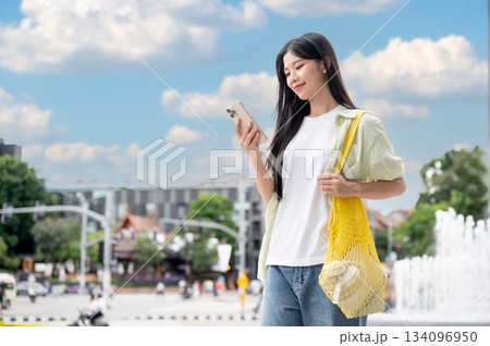 Asian woman looking at phone holding yellow net bag while standing or walking outdoor under blue sky Asian woman looking at phone holding yellow net bag while standing or walking outdoor under blue sky 134096950