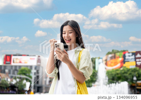 Smiling asian woman looking at smartphone and making a fist while standing outdoor under blue sky. 134096955