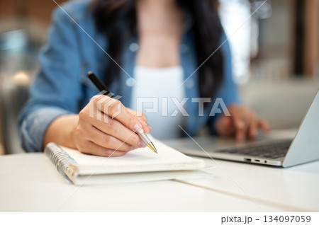 Close up woman student holding pen writing in notebook sitting in front of a laptop on table in cafe 134097059
