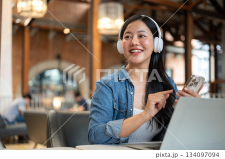 Happy asian woman wearing headphones looking away from phone and laptop sitting at table in the cafe Happy asian woman wearing headphones looking away from phone and laptop sitting at table in the cafe 134097094