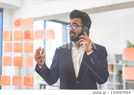 An Arab businessman in a suit talks on the phone while looking at sticky notes on a glass wall 134097664