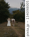 A girl in a white dress walks with horse on grassy path through trees at dusk in mountain area 134098005