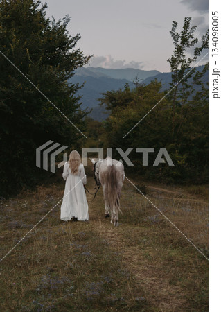 A girl in a white dress walks with horse on grassy path through trees at dusk in mountain area 134098005