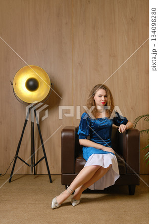 Woman sits in chair with styled hair and dress in indoor setting with light fixture Woman sits in chair with styled hair and dress in indoor setting with light fixture 134098280