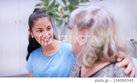 Adult granddaughter smiling while comforting her senior grandmother indoors Adult granddaughter smiling while comforting her senior grandmother indoors 134098345