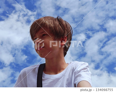 A boy looks upward with a contemplative expression under a bright blue sky with white clouds, wearing a black strap over his shoulder. Concept of wonder and daydreaming A boy looks upward with a contemplative expression under a bright blue sky with white clouds, wearing a black strap over his shoulder. Concept of wonder and daydreaming 134098755