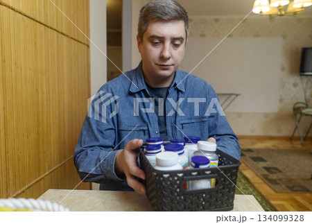 Man taking dietary supplements at home, pouring pills from a bottle and drinking with water. Concept of daily vitamins, health care and wellness routine. 134099328