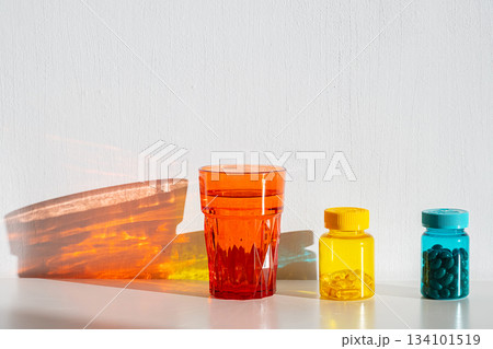 Colorful glass of water and pill bottles casting shadows on white wall. 134101519