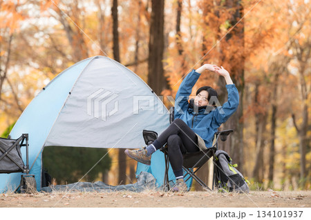 秋のキャンプで椅子に座りリラックスする男性ソロキャンパー、アウトドア楽しい～ストレッチで伸びる 134101937