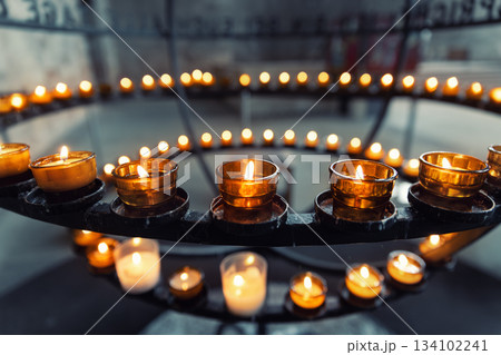 Burning votive candles warm glow in circular stand rack at French catholic cathedral church dark background. Prayer, hope, faith, remembrance prayer spirit in tranquil atmosphere Burning votive candles warm glow in circular stand rack at French catholic cathedral church dark background. Prayer, hope, faith, remembrance prayer spirit in tranquil atmosphere 134102241