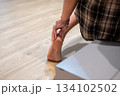 Relaxed Asian woman massaging her sore ankle near her bed, highlighting comfort and pain relief 134102502