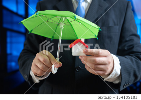 Concept of security and property insurance. Home safety, mortgage protection, real estate investment, family assets. Insurance agent holds an umbrella and a small house in his hands. Concept of security and property insurance. Home safety, mortgage protection, real estate investment, family assets. Insurance agent holds an umbrella and a small house in his hands. 134105183