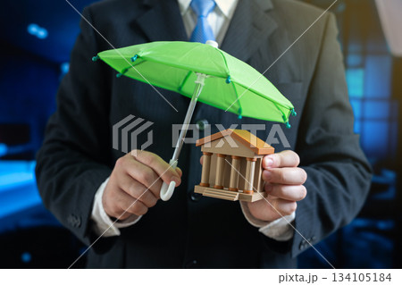 Businessman holds a green umbrella over a bank or government building. Protection of investments and financial well-being. Investment security, bank insurance, safe savings, and risk management Businessman holds a green umbrella over a bank or government building. Protection of investments and financial well-being. Investment security, bank insurance, safe savings, and risk management 134105184