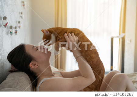 A brunette caucasian woman kisses and plays with a ginger domestic cat while sitting on a bed 134105265