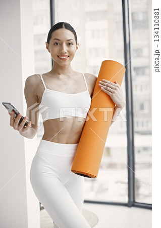 Slim and beautiful woman wearing white leggins and top and holding rolling yoga mat. Young girl with fit body posing in studio with big windows. Sport, fitness, diet, nutrition and health care concept Slim and beautiful woman wearing white leggins and top and holding rolling yoga mat. Young girl with fit body posing in studio with big windows. Sport, fitness, diet, nutrition and health care concept 134110881