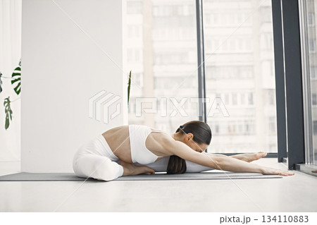 Slim and beautiful woman wearing white leggins and top and stretching on yoga mat. Young girl with fit body posing at bright studio with big windows. Sport, fitness, diet, nutrition and health care Slim and beautiful woman wearing white leggins and top and stretching on yoga mat. Young girl with fit body posing at bright studio with big windows. Sport, fitness, diet, nutrition and health care 134110883