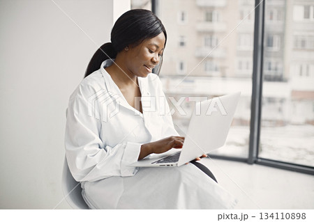 Young female black doctor sitting in her office at clinic near big windows. Woman wearing white medical coat. Brunette medical worker using a laptop. 134110898