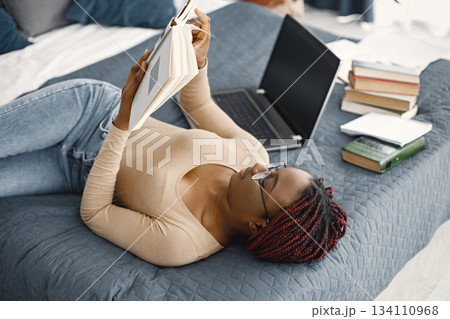 Young black woman lying down on bed reading a book at home. Girl wearing eyeglasses and beige sweater. Girl preparing her homework. 134110968