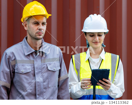 Two logistics workers wearing safety helmets and uniforms, standing by a shipping container. Female worker holding a tablet, managing tasks. Concept of teamwork, technology in logistics, and workforce Two logistics workers wearing safety helmets and uniforms, standing by a shipping container. Female worker holding a tablet, managing tasks. Concept of teamwork, technology in logistics, and workforce 134113362
