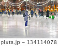 Woman ice skating outdoors on festive evening ice rink, surrounded by people under glowing string lights. Concept of winter joy, ice skating fun, seasonal leisure, urban Christmas atmosphere 134114078