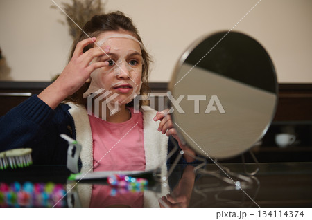 Elementary age Caucasian girl applying moisturizing face mask, siting at a table and admiring her appearance in a cosmetic mirror. The concept of home spa and skin care 134114374