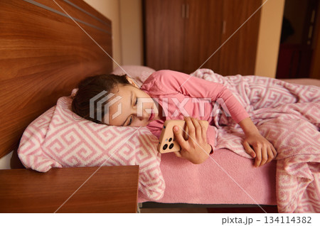 Child girl checking her mobile phone in the morning, still lying on her bed Child girl checking her mobile phone in the morning, still lying on her bed 134114382