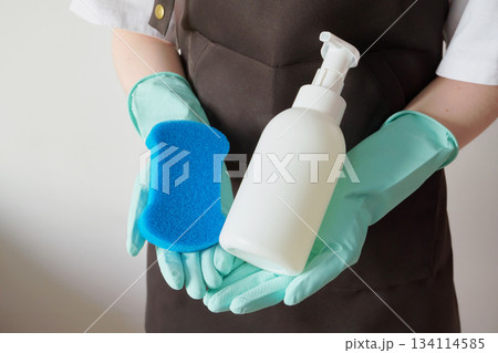 Woman wearing an apron and rubber gloves holding blue sponge and white soap dispenser. Woman wearing an apron and rubber gloves holding blue sponge and white soap dispenser. 134114585