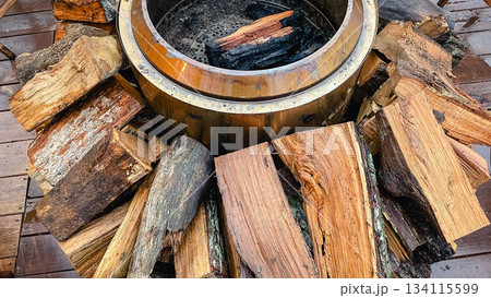 Close up of a warm metallic fire pit surrounded by chopped firewood and wet wooden deck boards 134115599