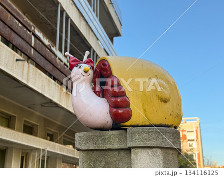 Playful snail sculpture with bow standing on concrete pedestal under blue sky. Whimsical street art, vivid decoration, joyful urban detail bringing charm and character to city space. Playful snail sculpture with bow standing on concrete pedestal under blue sky. Whimsical street art, vivid decoration, joyful urban detail bringing charm and character to city space. 134116125