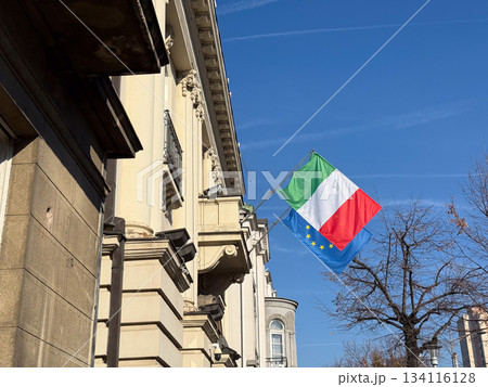 Italian and European flags waving near historic building under clear sky. Symbol of cultural connection, diplomacy, political unity and national identity in urban environment. 134116128