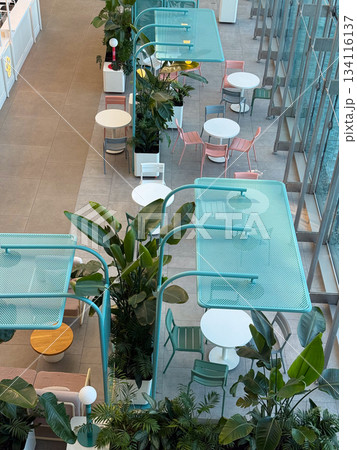 Cafe seating area with plants and pastel chairs under blue mesh shades. Social interaction, leisure, modern hospitality space, relaxed atmosphere and stylish public interior. 134116137