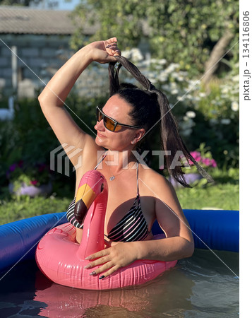 Woman wearing sunglasses is holding her hair and relaxing on inflatable flamingo in swimming pool Woman wearing sunglasses is holding her hair and relaxing on inflatable flamingo in swimming pool 134116806