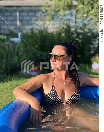 Woman relaxing in inflatable pool in garden during summer Woman relaxing in inflatable pool in garden during summer 134116808