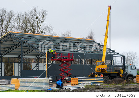 Workers installing sandwich panels on steel frame building using lift and crane at construction site 134117658
