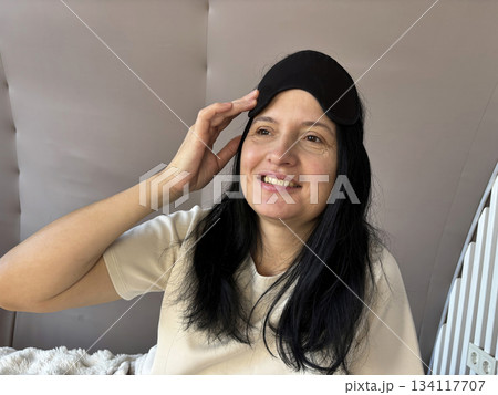 Adult brunette woman sitting in bed with a sleep mask, smiling and looking relaxed after a good night sleep, cozy morning routine, healthy sleep, wellness and self care lifestyle 134117707