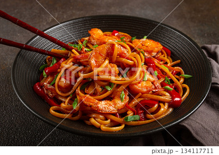 udon stir-fry shrimp noodles with red bell pepper and green onion, served in a black bowl, with chopsticks, dark background, 134117711