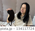 Adult brunette woman sitting in bed with a sleep mask, smiling and looking relaxed after a good night sleep, cozy morning routine, healthy sleep, wellness and self care lifestyle 134117724
