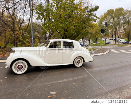 Vintage white car driving along autumn road. Retro style, nostalgia, classic transport and elegant automotive heritage Vintage white car driving along autumn road. Retro style, nostalgia, classic transport and elegant automotive heritage 134118312